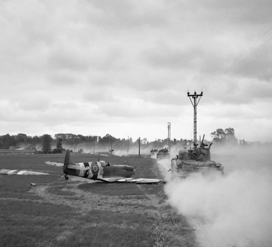 Sherman tanks move up past a crash landed Spitfire, for an attack on Tilly-sur-Seulles, Normandy 17 June 1944
