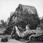 Soldier reads newspaper by camouflaged M7 near Carentan, 1944